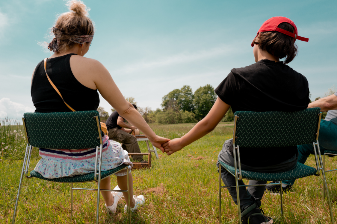 A group of people sitting in chairs set up in a circle outside. The focus is on two people, facing away from the camera, holding hands.