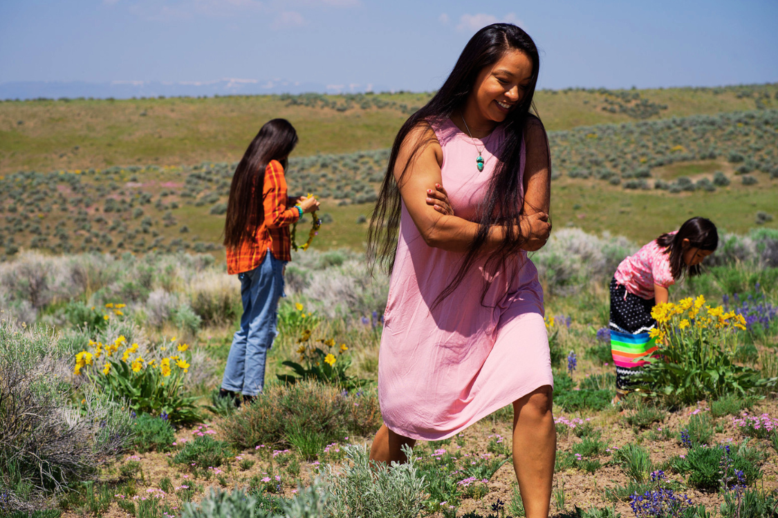 In an arid grassland on a sunny day, three Native people, one adult in a pink sleeveless dress and two children, one in a pink top and colorful ribbon skirt and the other in a jeans and an orange shirt, walk through the foreground and pick yellow flowers from among the growing plants.